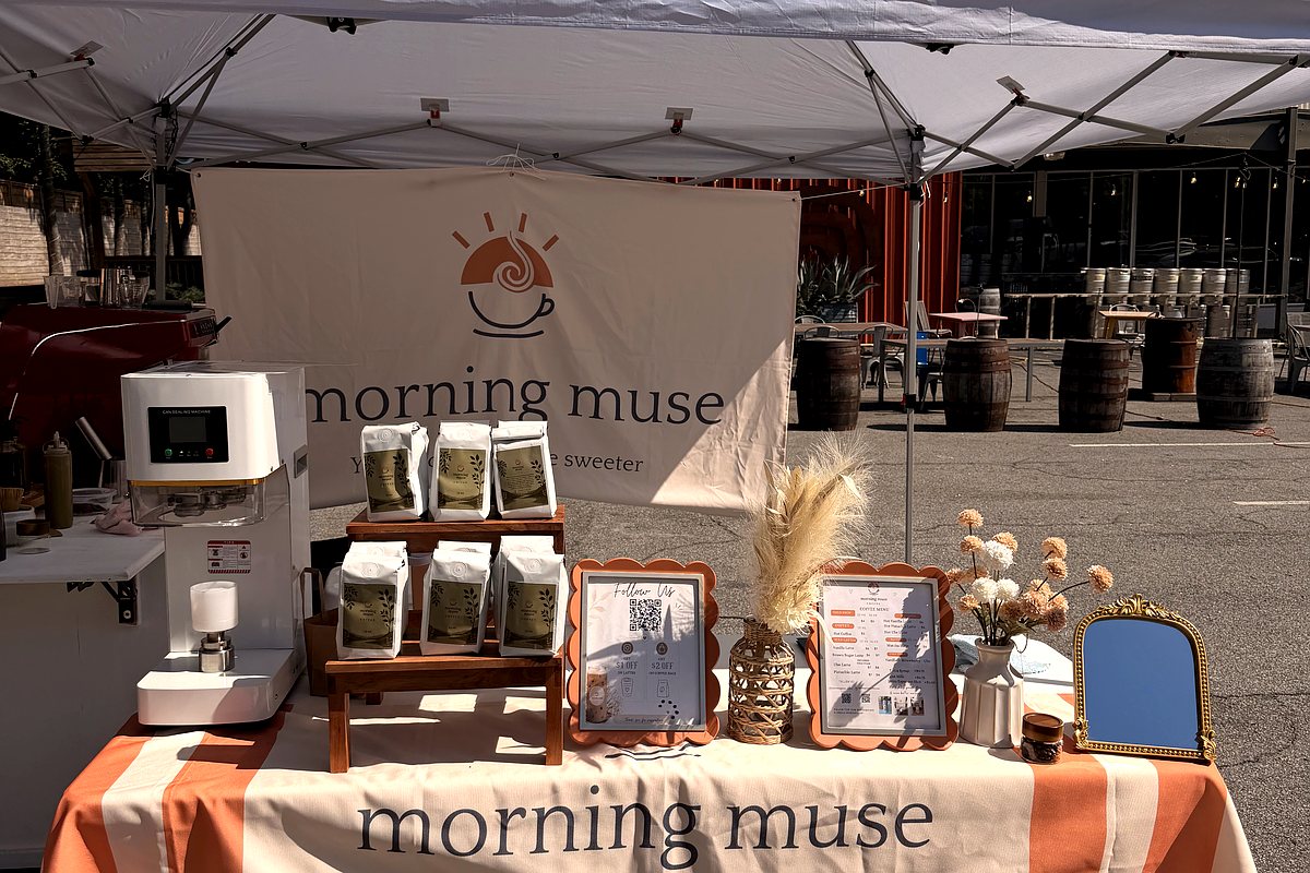 The Morning Muse pop-up table at the market: banner, coffee bags on display, and the menu