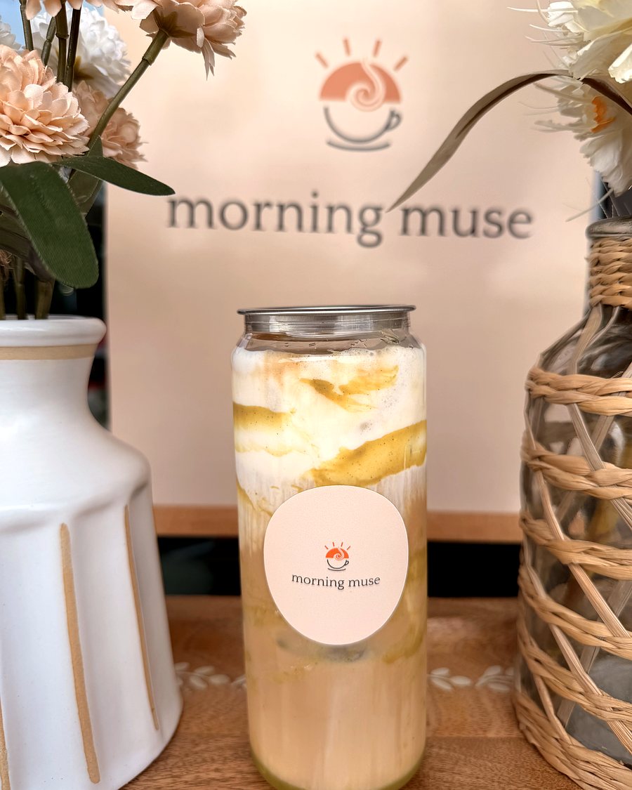 Pistachio latte in a Morning Muse can, soft green with a caramel swirl, on the branded backdrop